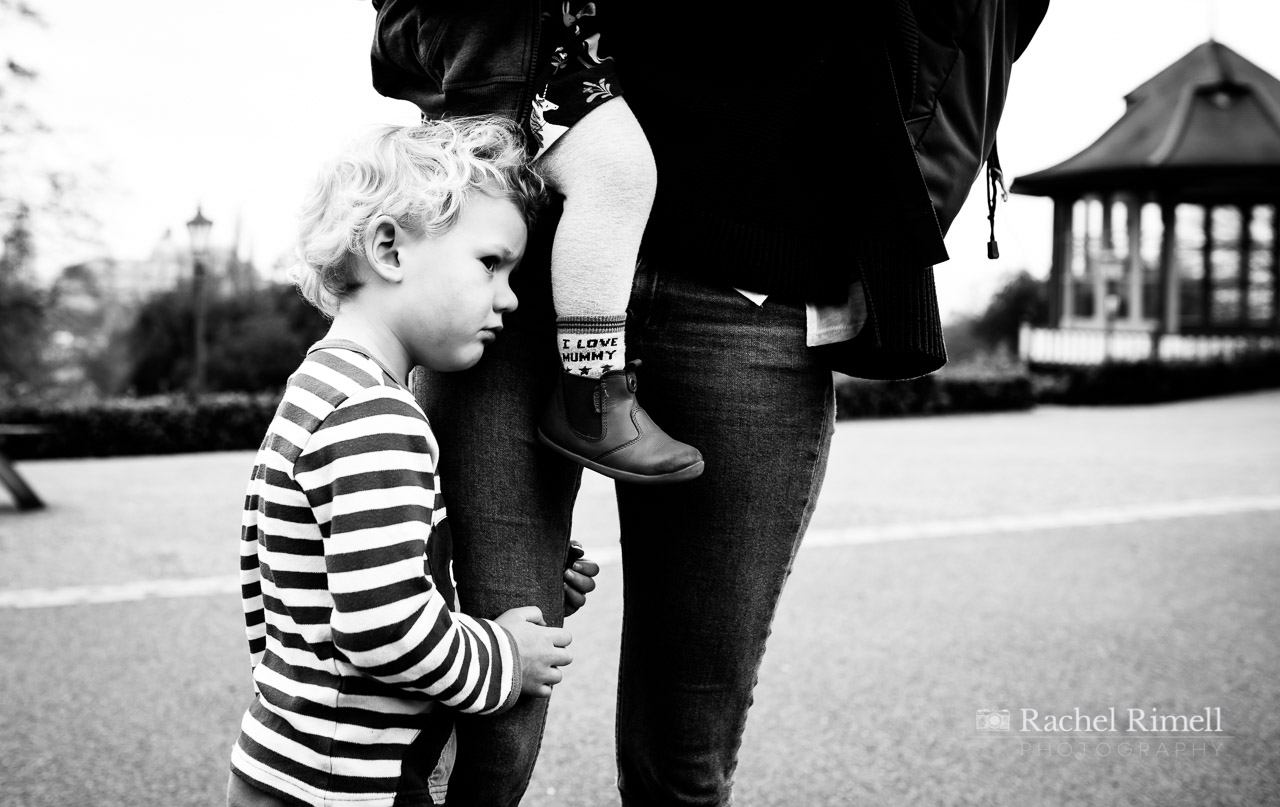 son holds onto mum's leg. Mum holds younger daughter with I love mummy socks. Horniman Gardens