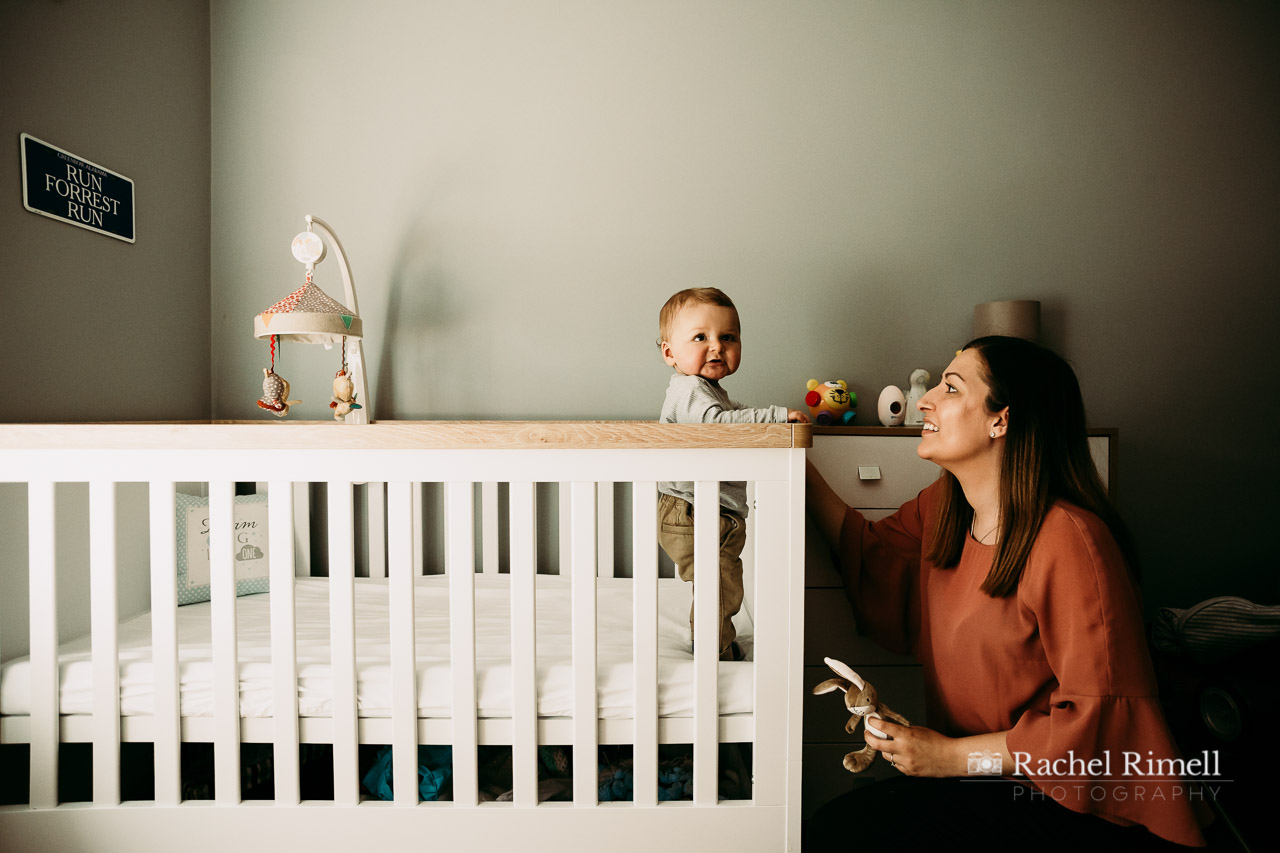 Mother looks at son standing in cot documentary child portrait