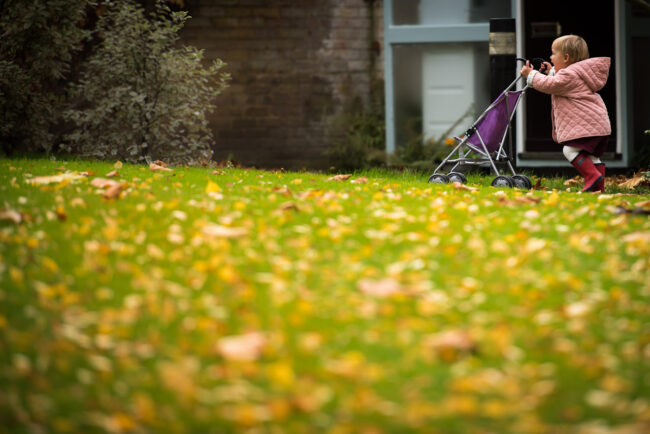 toddler girl in pink jacket and red wellies running with a purple toy pram in Blackheath on a natural family photography session
