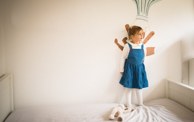 Young girl in a blue dress standing on her bed with white wall behind and picture of fox on a hot air balloon partially obscured, Blackheath fun family photography
