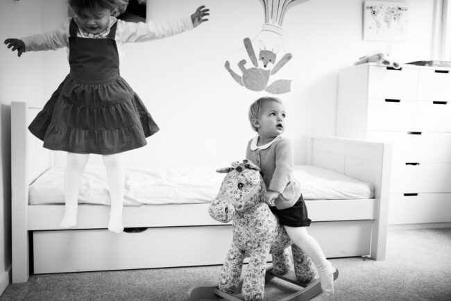 black and white image of girl jumping off her bed while younger sister looks off camera riding a rocking horse with image of fox on a hot air balloon in background as part of a day in the life documentary family photography session UK