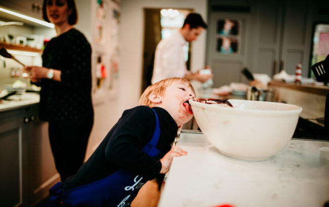 Boy licking bowl after making brownies in kitchen with mum and dad in the background on a relaxed day in the life documentary family photography session in Clapham