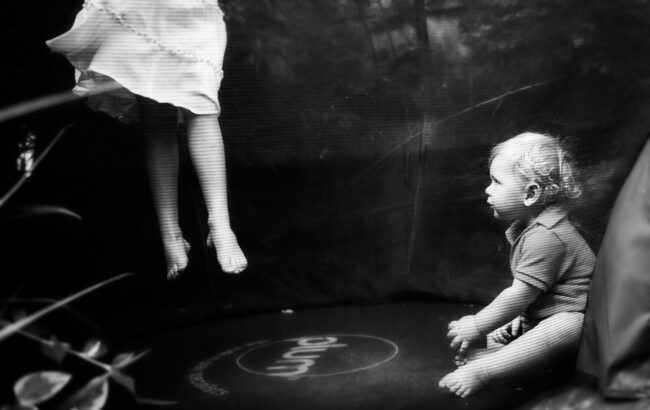 fun family photo of girl on a trampoline with her legs showing lifting off with her young brother looking on. Taken in Forest Hill.