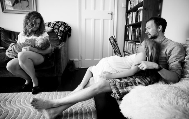 unconventional family portrait at home in London with father and daughter ready on sofa and mum breastfeeding son