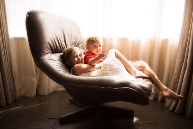 girl and toddler brother playing on a large mid-century modern brown swivel chair. Boy wearing red t-shirt and girl wearing a white summer dress. With curtains and window behind. Natural children photography London