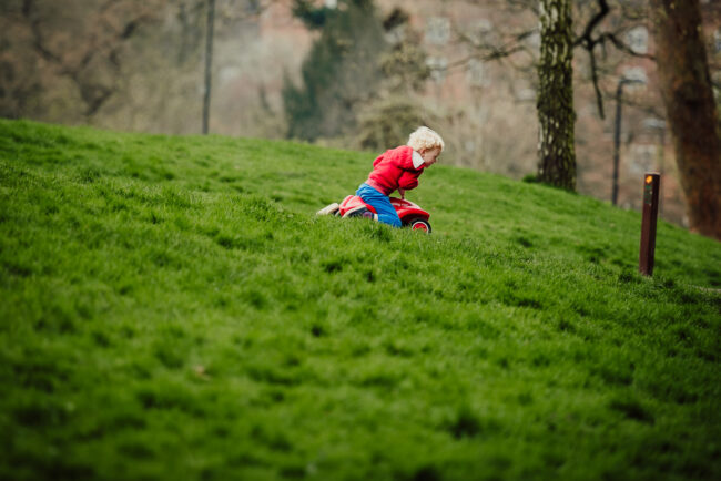 boy in red top and blue trousers racing red ride on car down a hill in relaxed family photography session outdoors in London