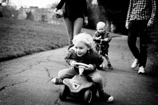 alternative family portraits boy racing on his ride on car with sister on scooter in the background and parents legs visible. Black and white photo. London