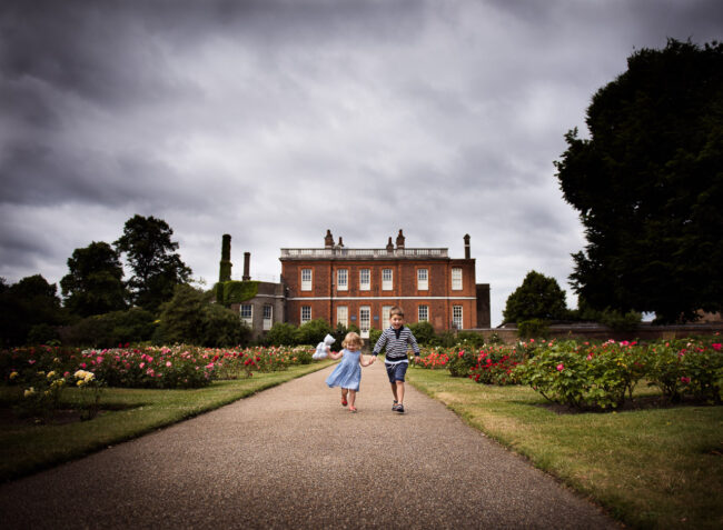 Brither and sister run towards the camera with The Rangers House, Greenwich Park in the background with cloudy sky above on a fun outdoor family photography, Greenwich Park