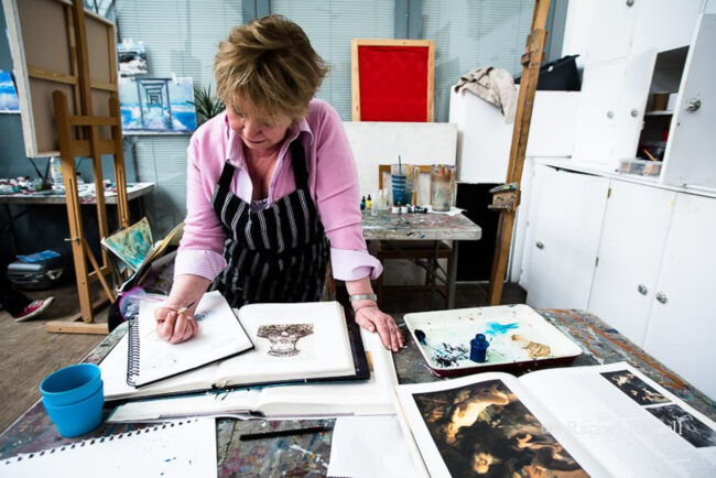 environmental portrait of artist at work sketching with art books strewn about and easel in background, London