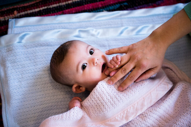 baby looking at camera under a pink blanket holding mum's finger to her mouth in Streatham natural baby portrait