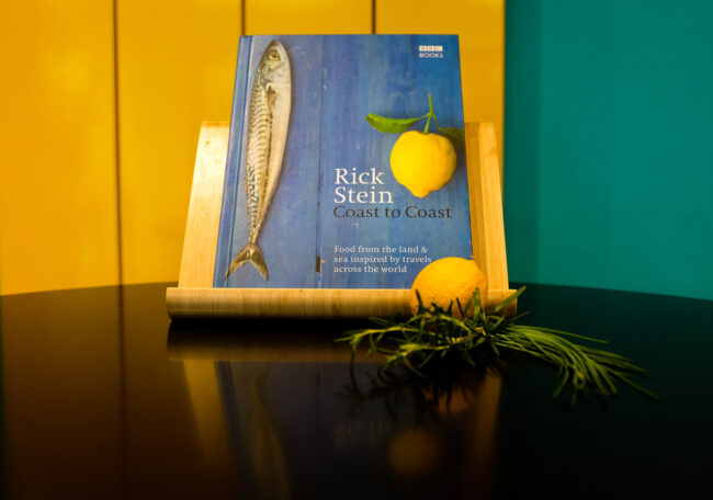 creative food and product photo of Rick Stein recipe book in blue with lemon on cover on a black table with a lemon and herbs, against a yellow and turquoise background, London