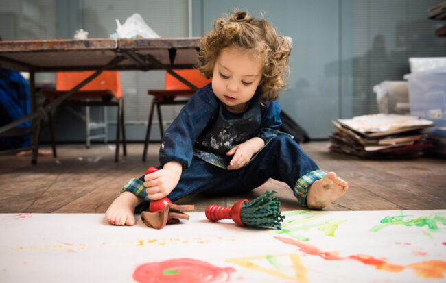 toddler-aged girl sitting on a wooden floor painting a large strip of paper with a sponge, wearing a painting apron at a child art class at Blackheath Conservatoire