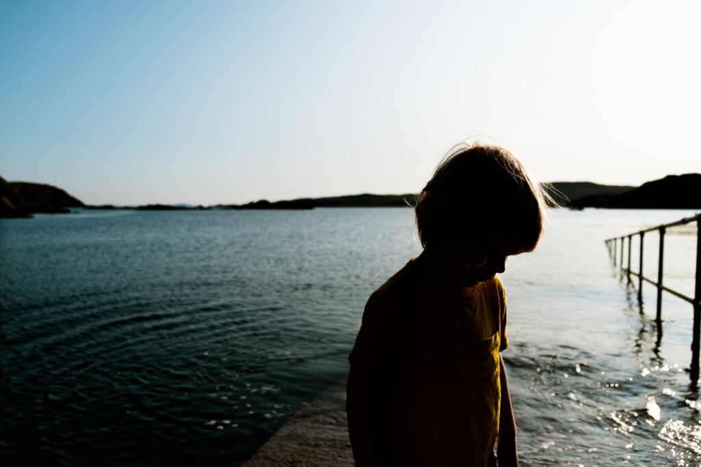 candid alternative portrait of boy in yellow t-shirt with profile in shade and the sea behind. UK vacation photography