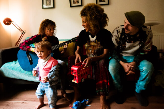 Funa nd relaxed family portrait sitting on their sofa in South London while eldest daughter plays blue guitar. Young toddler boy wandering out of frame while mother wearing a lion t-shirt and vibrant trousers holds a toy bus and father in a flat cap, animal jumper and blue jeans looks on.