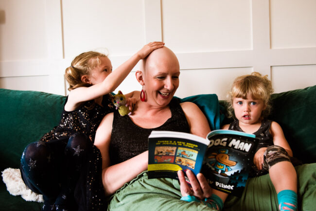 mum with bald head during chemotherapy cancer treatment wearing big dangly red earings reading a story to her two daughters on a green sofa while elder girl strokes her bald head on a relaxed documentary-style family photography session