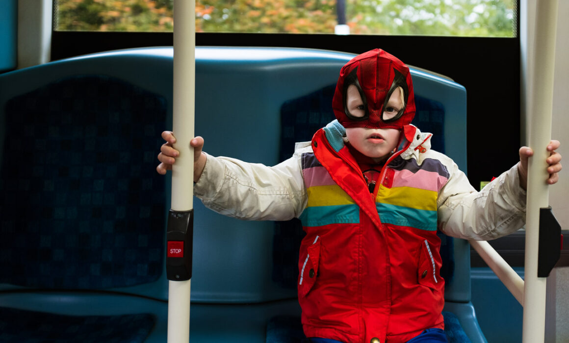 young boy with a spiderman facemask on and colourful winter jacket and a grumpy face on the London bus in a natural child portrait in London, UK