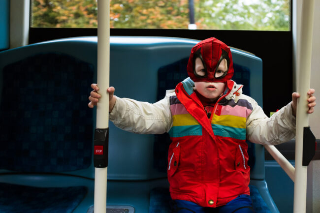 young boy with a spiderman facemask on and colourful winter jacket and a grumpy face on the London bus in a natural child portrait in London, UK