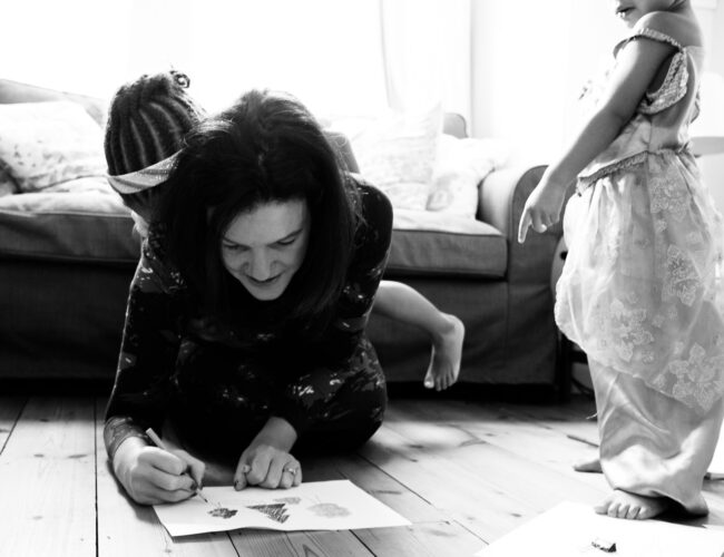 black and white photo of mum drawing with two daughters climbing on top of her on a natural unposed family photography session Sydenham south London