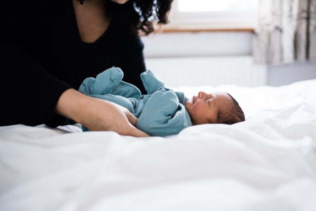 newborn bab in blue babygro on the bed with mother's arms in real life natural newborn photography session London