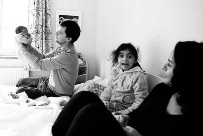 Forest Hill newborn photography session with dad holding newborn baby up, while older sister pulls a face at the baby crying while mother looks on in black and white