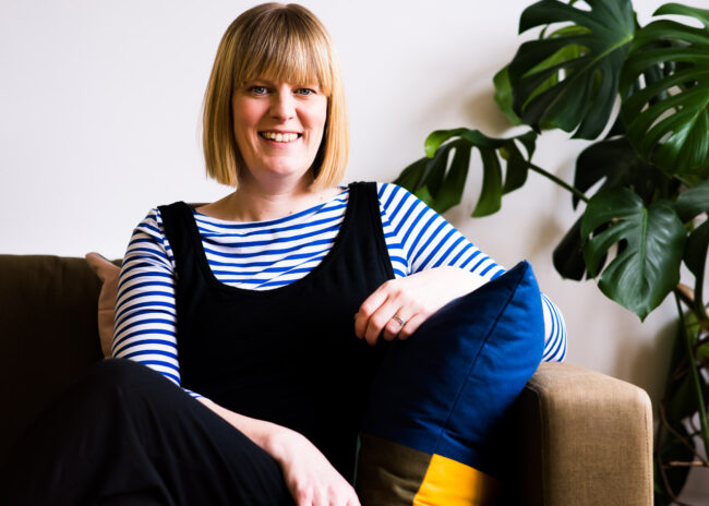 relaxed business headshot of life coach Janine Green on brown sofa with blue and yellow cushion, wearing striped top and black dungarees with green cheese plant in background, Crystal Palace, London