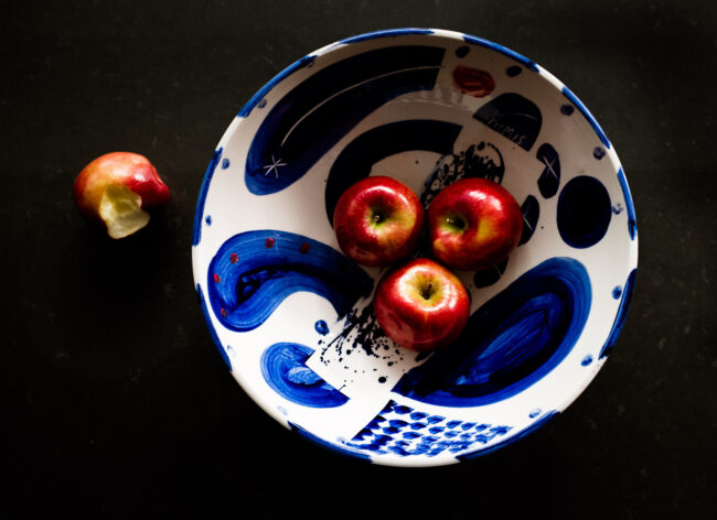 stylised food photo of red shiny apples in a blue and white patterned bowl with an apple with a bit missing outside the bowl on a black background, London