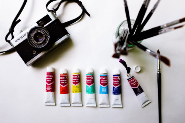 flat lay of paints in rainbow colours with jar of paintbrushes and vintage camera, london