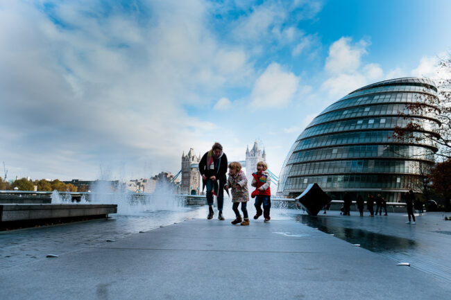 London vacation photography family running away from the fountains at More London with City hall and Tower Bridge in background