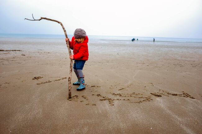 Girl in bright red coat, woolen hat, denim dress and striped wellies drawns in the sand with a large branch at Harlech Beach in Wales. Alternative family vacation photography UK