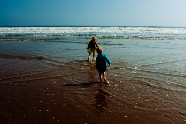 Brother and sister running into the waves wearing their clothes (yellow dress and blue cardigan) at a beach in Northumberland. UK vacation photographer.
