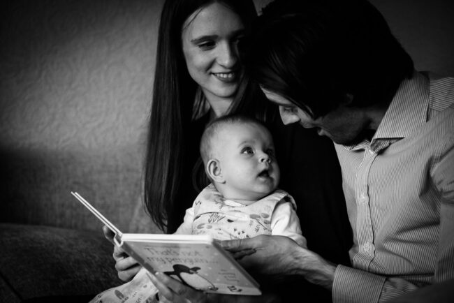 A natural relaxed family portrait of a mother and father reading to their baby daughter at their home in Orpington with baby looking up in awe at her dad, dad looking animated and mum smiling.