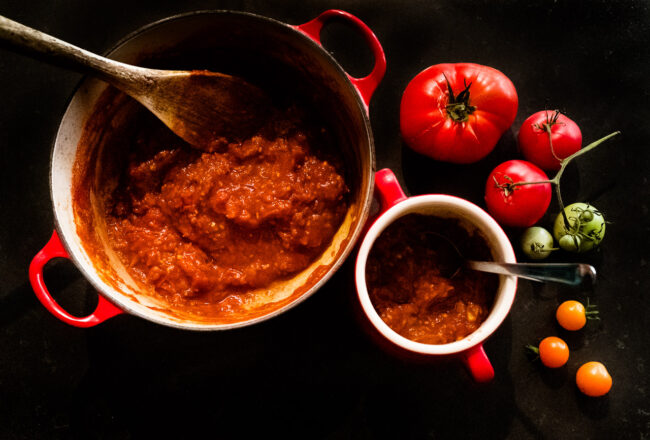 styled food photo of red and green and orange tomatoes and a large red pot with tomato chutney