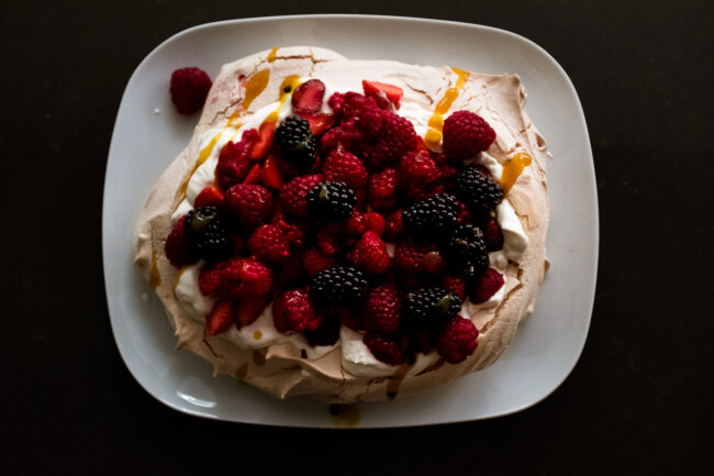 style food photo of a merangue with berries and mago coulis on a white plate on a granite background