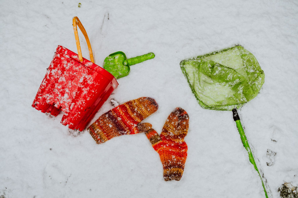 red child's bucket and green spade with green fishing net lying in the snow alongside a pair of orange woollen mittens