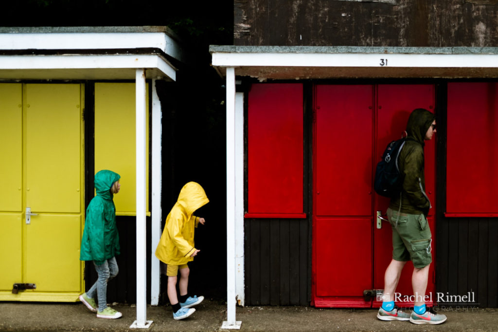 A dad with two children in the rain walking along the beach huts at Filey promenade wearing green and yellow raincoats