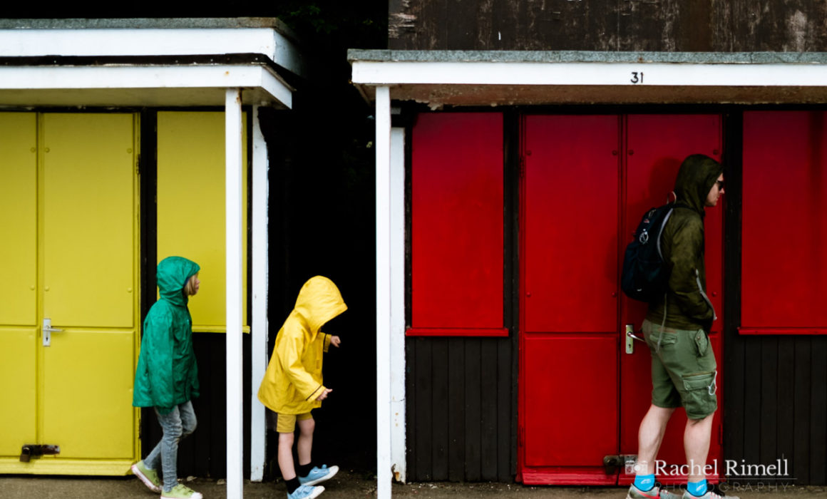 A dad with two children in the rain walking along the beach huts at Filey promenade wearing green and yellow raincoats