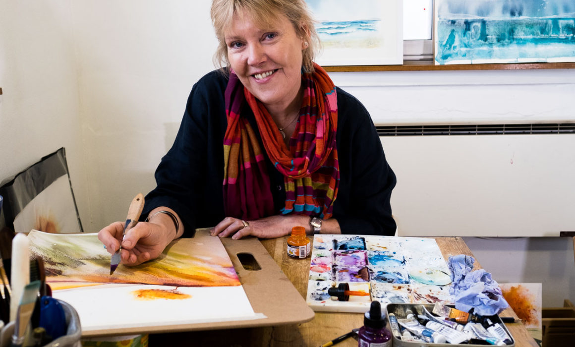 Watercolour artist Ione Harrison at her desk looking towards the camera and smiling with red scarf