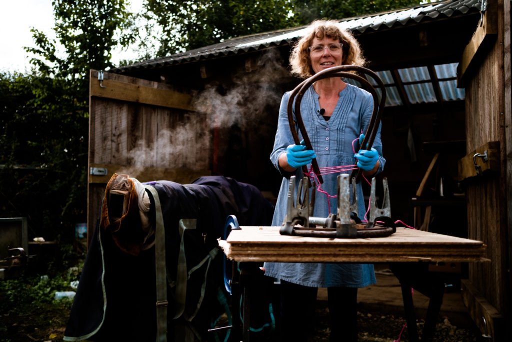 white woman in blue dress standing outside her workshop with two bent pieces of willow with her willow steamer in background