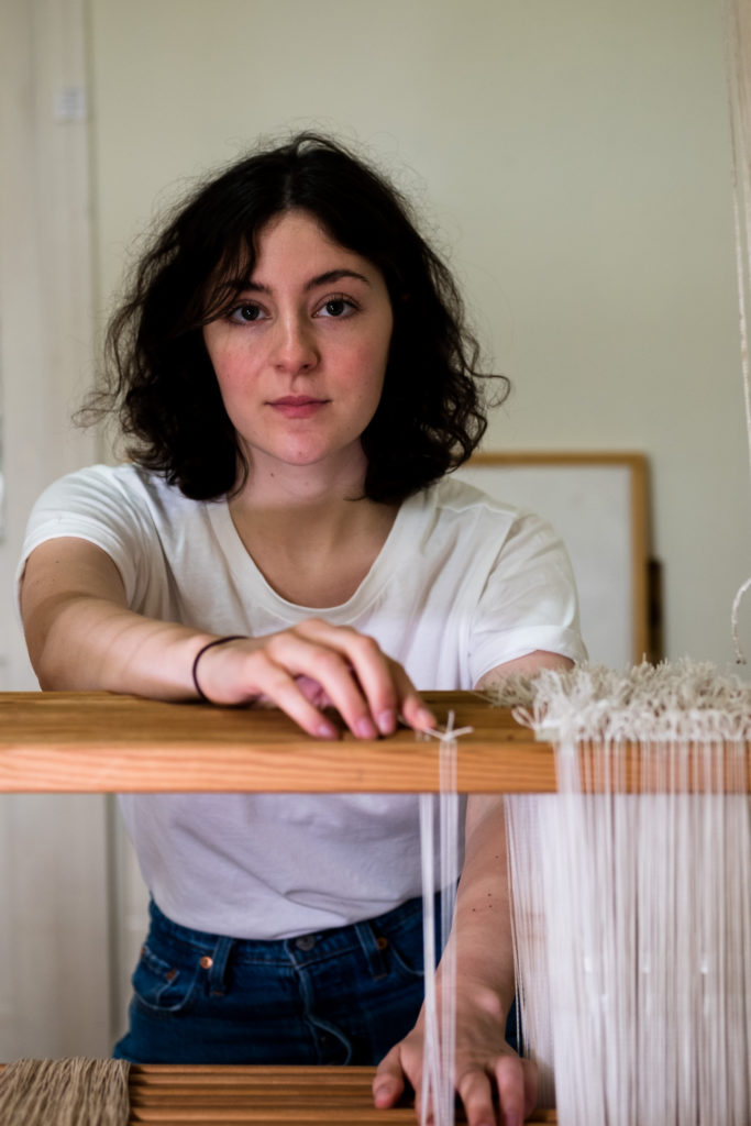 A young woman in a white t-shirt with dark curly hair standing at a traditional style loom