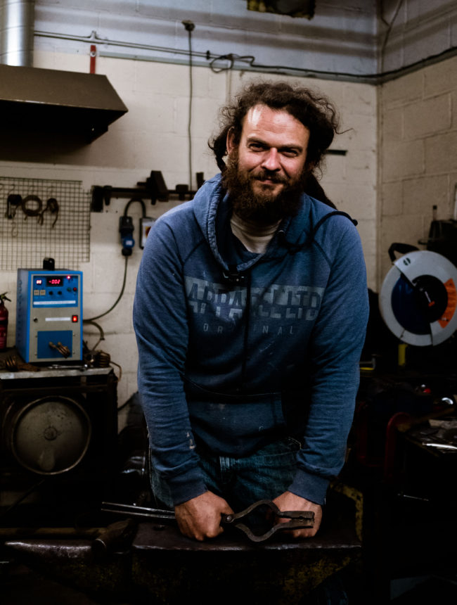 white male blacksmith with long dark dreadlocked hair and blue sweatshirt standing at an anvil holding a hammer