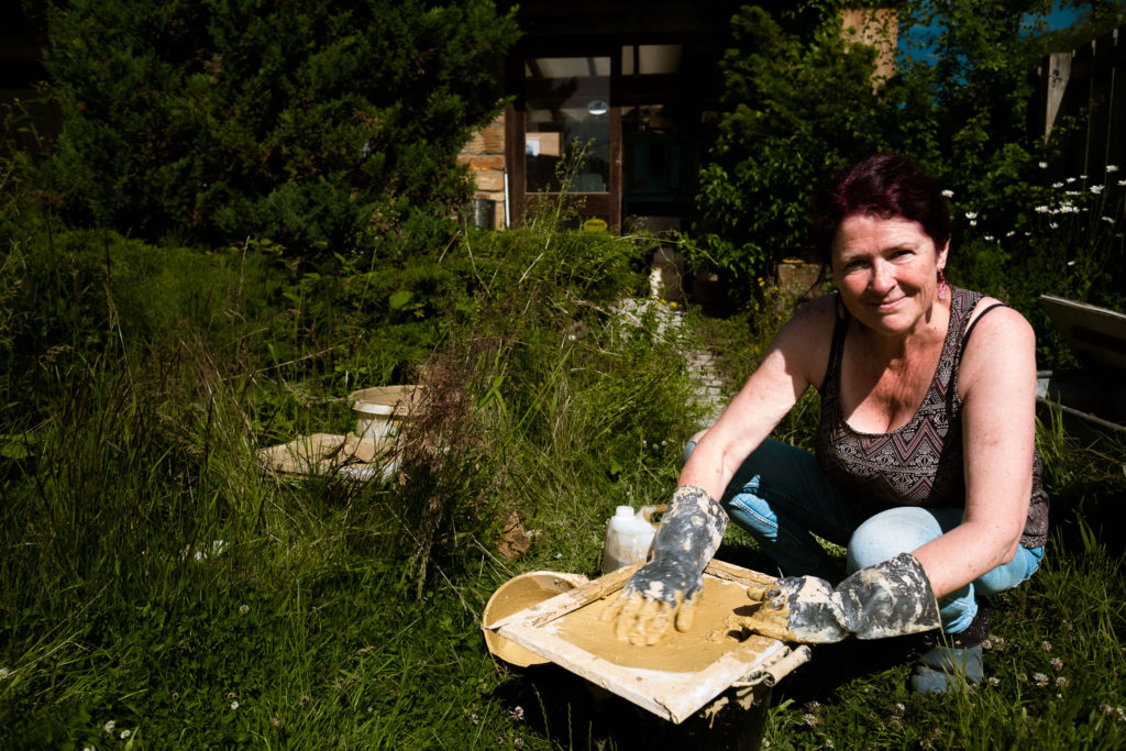 woman with long purple and dark hair crouched outside her pottery studio mixing wet clay on a screenprinting board as a sieve over a bucket, wearing rubber gloves