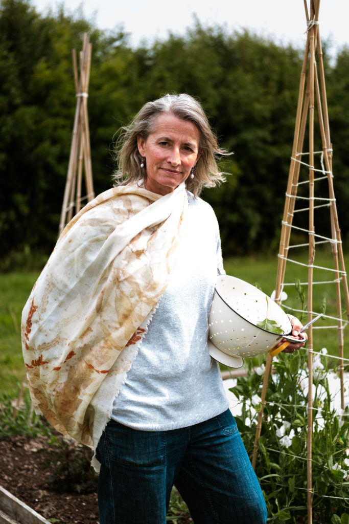 A white middle aged woman in a pale jumper with a botanical print chiffon scarf and shoulder length grey hair stands with a colander and a pair of scissors beside some pea climber frames in her kitchen garden