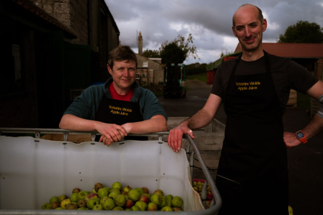 Woman with short dark hair and bald man, both in aprons, standing in front of a large container with apples with farm in background