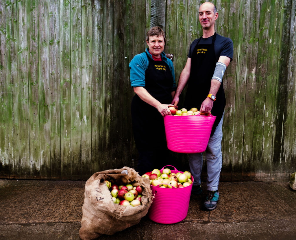 a white man and woman stand outside a green wooden shed with pink buckets and a sack filled with apples