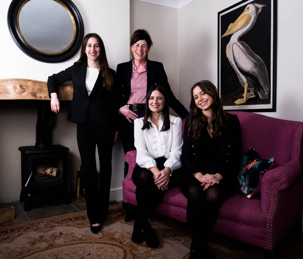 two white women with dark long hair on a pink sofa with two white women with long dark hair standing behind against a fireplace with large pelican painting on the wall, professional team photo