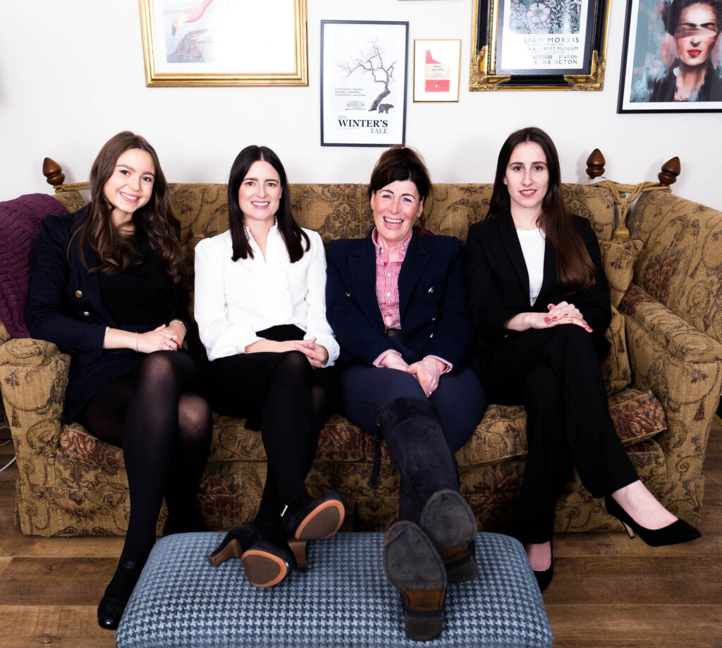 informal team photo of 4 white women sitting and laughing on a sofa with framed wall art behind